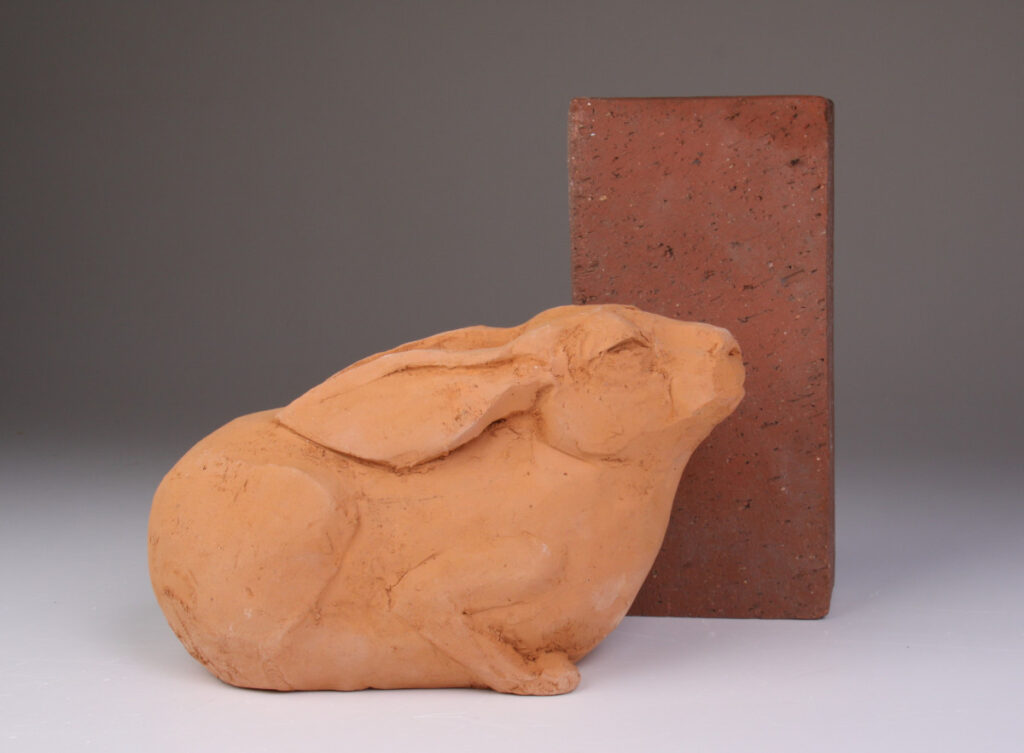 Keramikskulptur av en vilande hare bredvid en tegelsten. Gjord i röd lera från Wanås Gods.